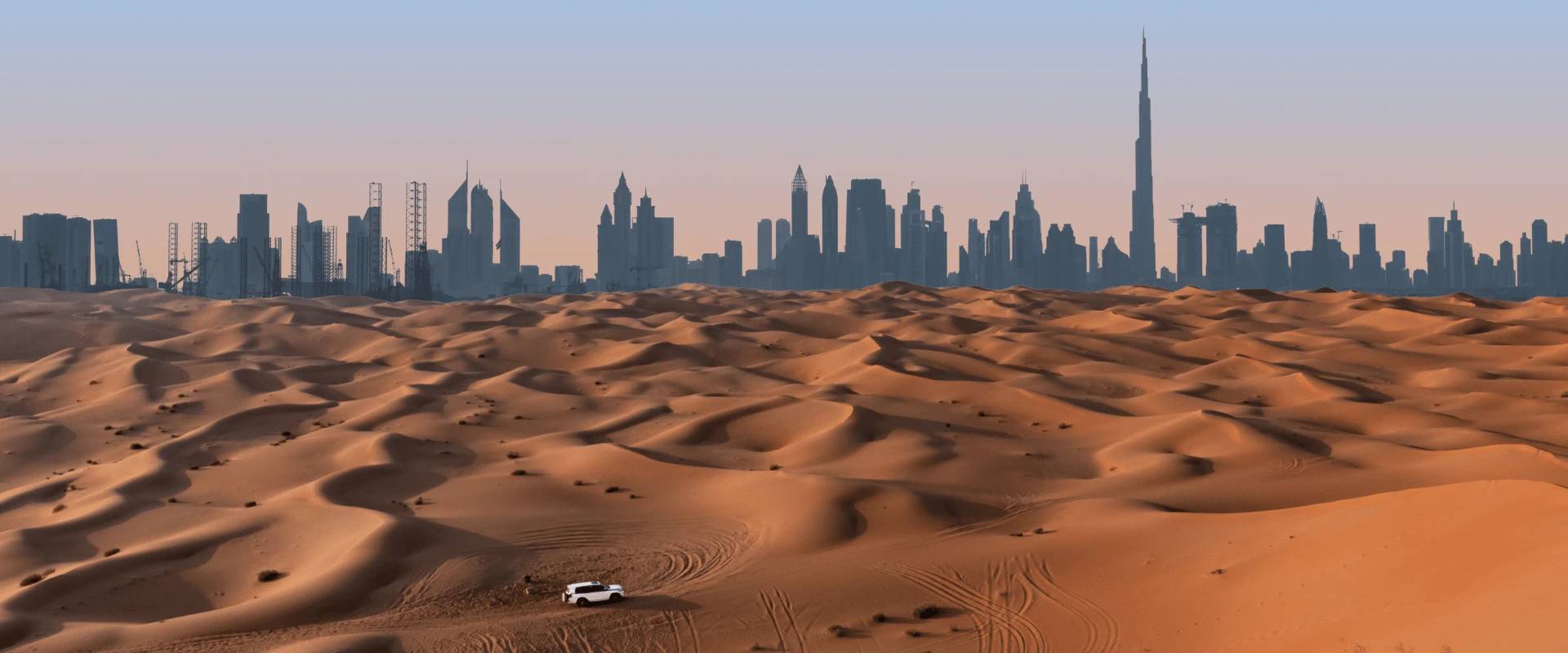 A white 4x4 on red sand dunes with the Dubai skyline and Burj Khalifa on the horizon