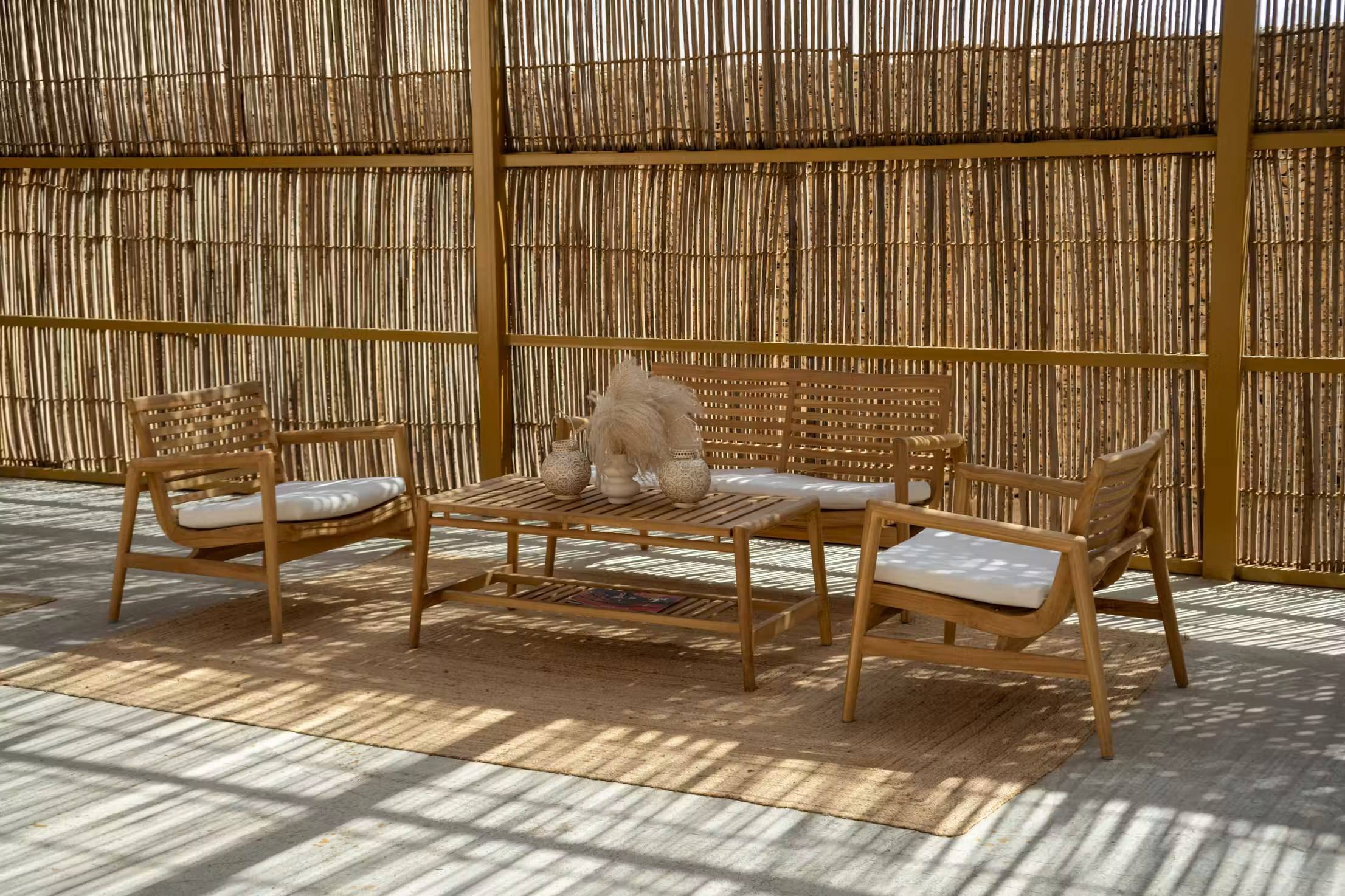 Wooden chairs and a low table in a shaded seating area with palm-frond walls