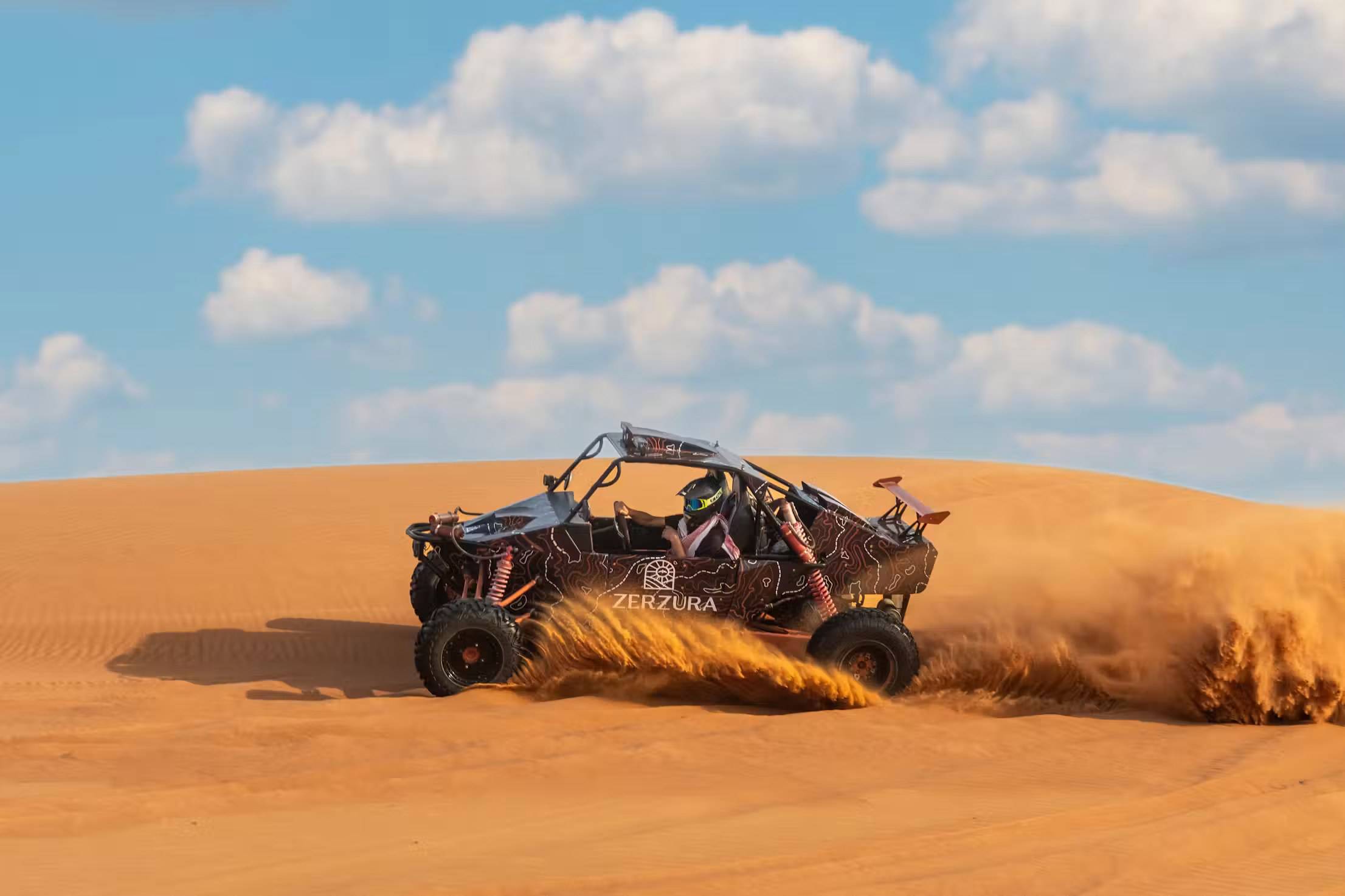 A dune buggy kicking up sand on a dune under a blue sky with clouds