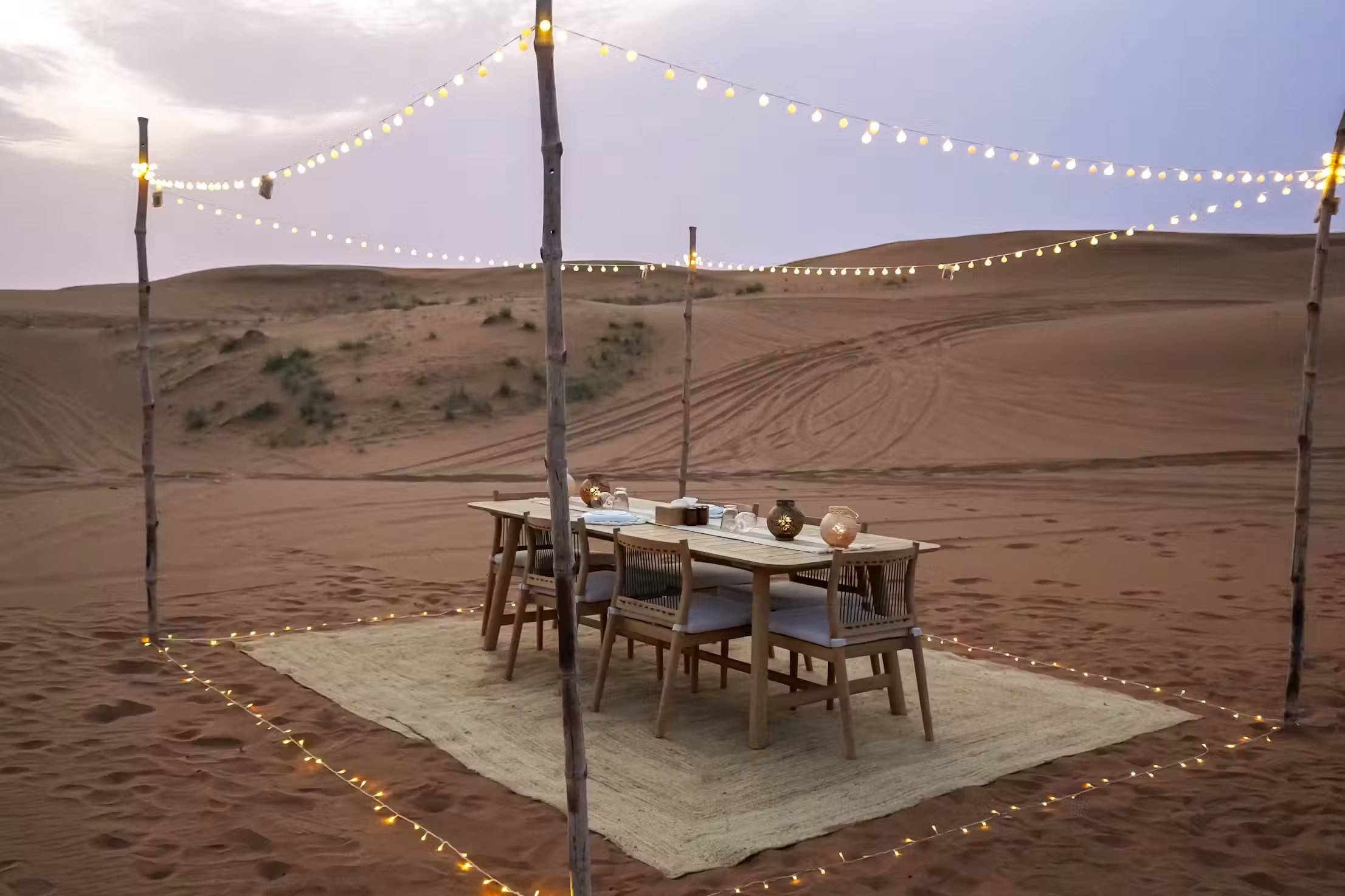 A dinner table set on a rug in the dunes under strings of lights